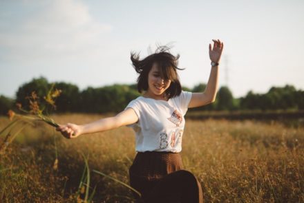 Kostenfreie geführte Selbstliebe Meditation - Bildbeschreibung: Eine junge Frau tanzt lächelnd in einem Feld voller hüfthoher Gräser.