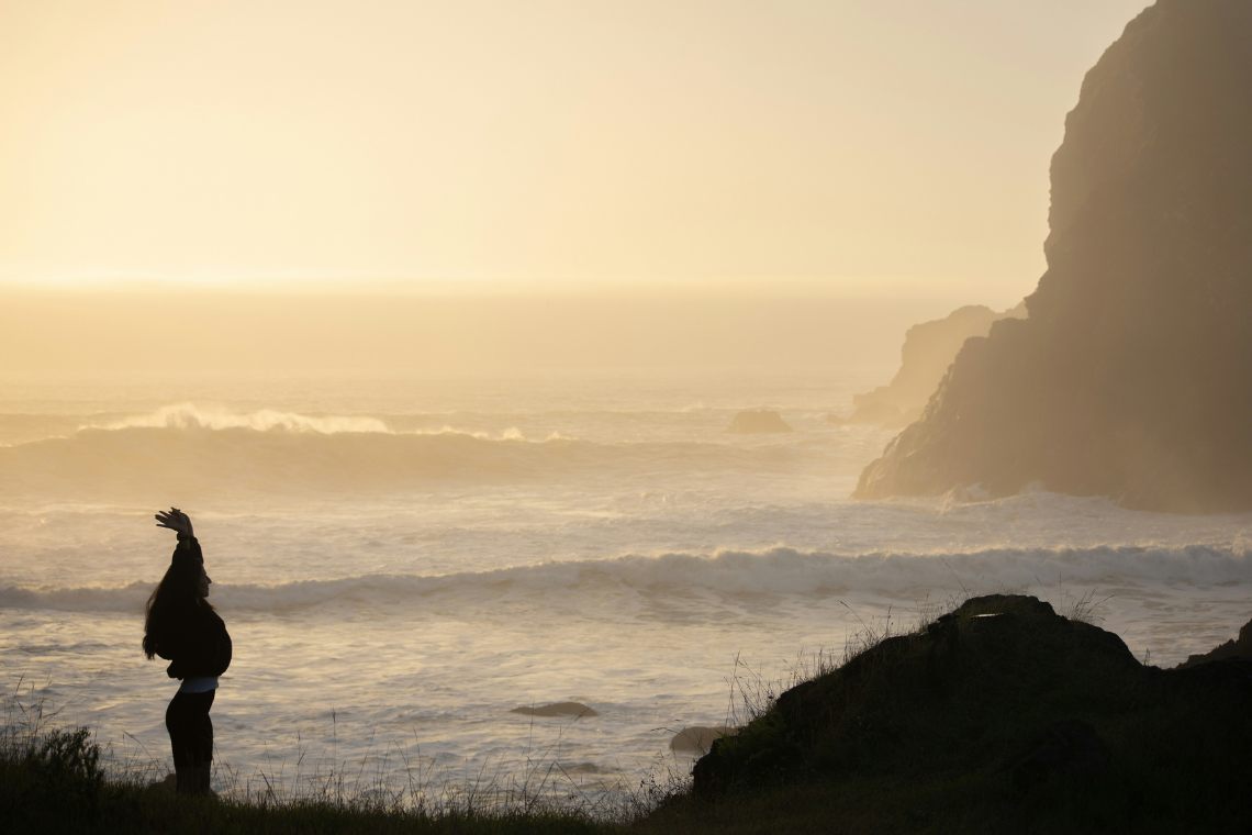 Beitragsbild zum Blog-Beitrag "Spirituelle Gaben – wie Du Dein Vertrauen in Deine Fähigkeiten stärkst". Auf dem Bild sieht man eine Frau, die auf einer kleinen Klippe am Meer steht und die Arme in die Höhe streckt. Am rechten Bildrand sieht man einen großen Felsen oder eine Klippe. Das Licht ist weich und die Luft ist diesig. Es ist starker Wellengang. Es wirkt wie ein morgendliches oder abendliches Licht.