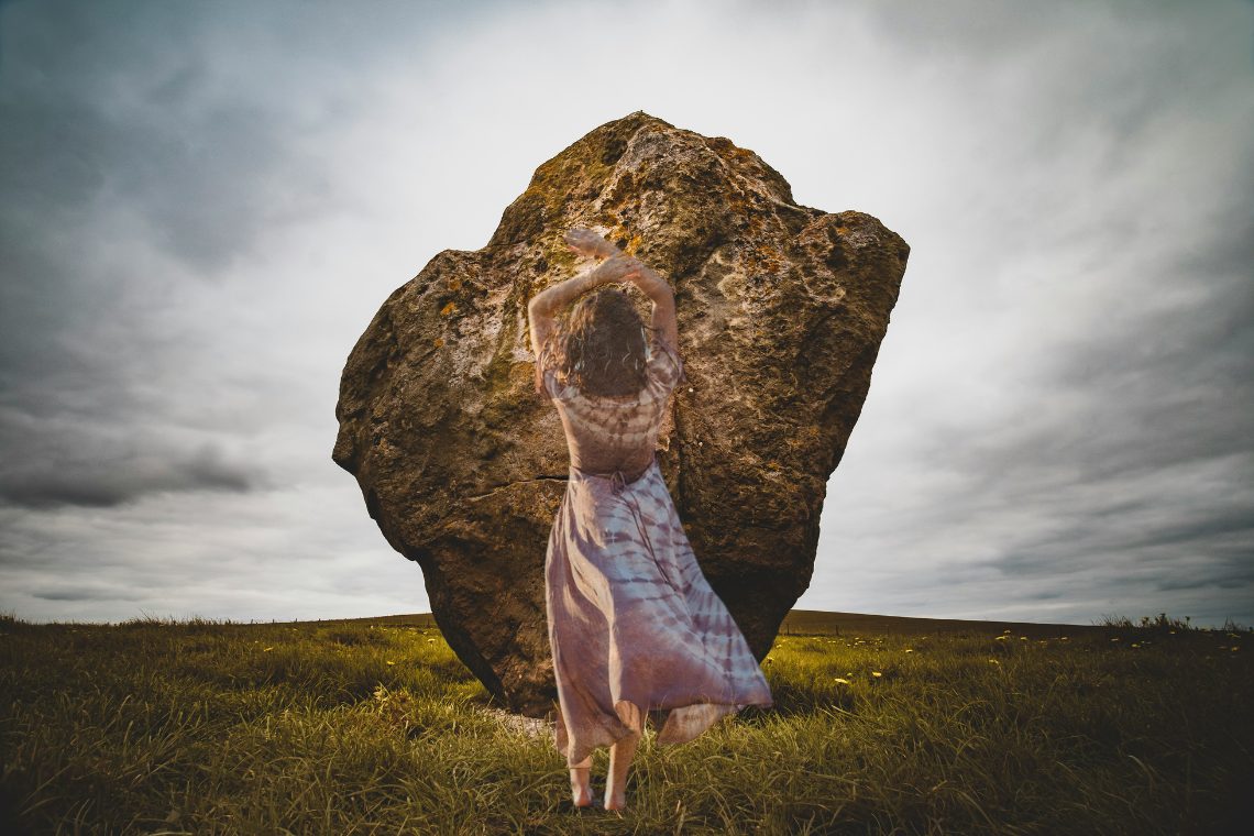 Ideen für Imbolc Rituale von Suzanne Frankenfeld [Bildbeschreibung: Ein großer Stein steht auf einer grünen Wiese vor bewölktem Himmel. Aufgenommen in Avebury, UK. In das Bild hineinmontiert ist eine von hinten fotografierte Frau mit wallendem Kleid.]