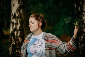 Beitragsbild zur Podcastfolge "Im Zwischenland mit Skunk Anansie" von Suzanne Frankenfeld. Auf dem Bild zu sehen ist Suzanne, wie sie in einem Waldstück steht. Ihre Augen sind geschlossen und sie berührt mit der linken Hand eine Birke.