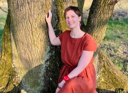Jenny Festerling, Fachärztin für Allgemeinmedizin und Zyklusmentorin, spricht über Endometriose und Zyklusbewusstsein. Bildbeschreibung: Eine Frau in einem roten Kleid sitzt auf dem Stamm eines großen Baumes und berührt den Baum mit einer Hand. Sanftes Licht fällt auf ihr Gesicht. Sie lächelt zufrieden.