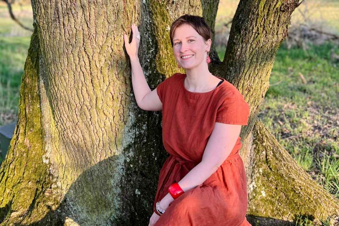 Jenny Festerling, Fachärztin für Allgemeinmedizin und Zyklusmentorin, spricht über Endometriose und Zyklusbewusstsein. Bildbeschreibung: Eine Frau in einem roten Kleid sitzt auf dem Stamm eines großen Baumes und berührt den Baum mit einer Hand. Sanftes Licht fällt auf ihr Gesicht. Sie lächelt zufrieden.